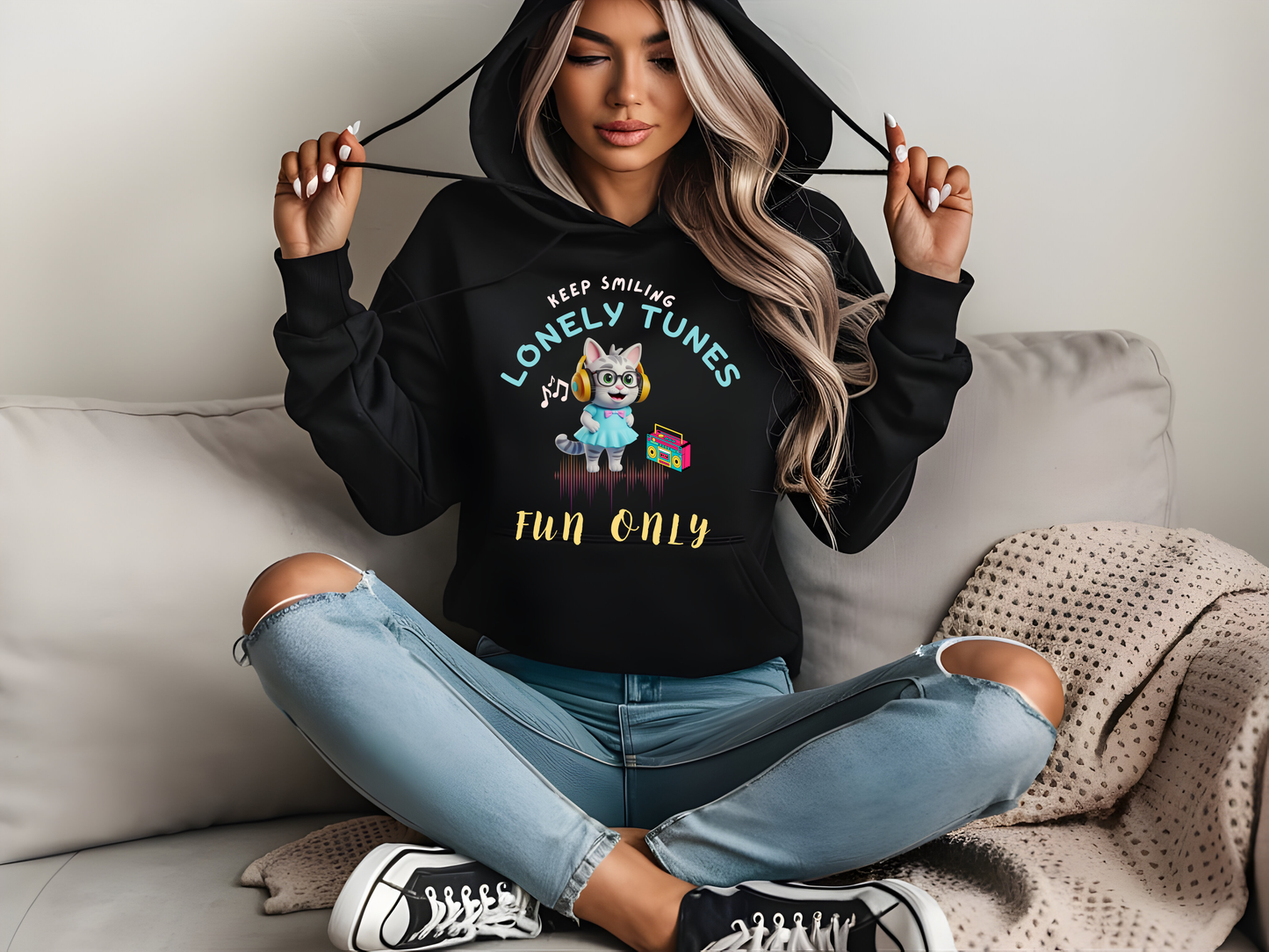 Black hoodie featuring a vibrant 'Lonely Tunes' cartoon character and music-themed design, worn by a woman sitting on a sofa in casual jeans.
