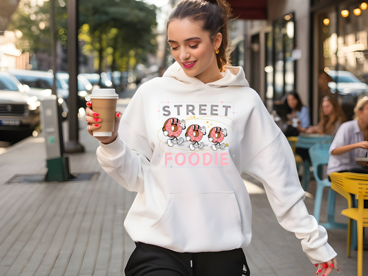 A stylish young woman wearing a white "Street Foodie" hoodie featuring playful doughnut graphics, holding a coffee cup while walking on a vibrant city street. The hoodie is perfect for casual outings and street style. The background showcases cafes and pedestrians, highlighting a lively urban atmosphere. Ideal for fashion-forward food lovers and trendy streetwear enthusiasts.