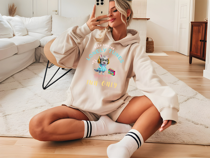 Beige hoodie featuring a playful 'Lonely Tunes' design with a cartoon character and retro music theme, worn by a woman sitting casually in a cozy living room.