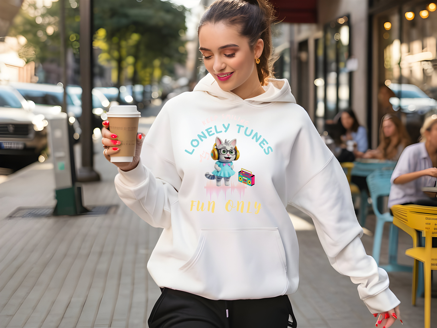 Stylish white hoodie with a colorful 'Lonely Tunes' design featuring a cheerful cartoon character and retro music elements, worn by a woman holding a coffee cup while strolling outdoors.