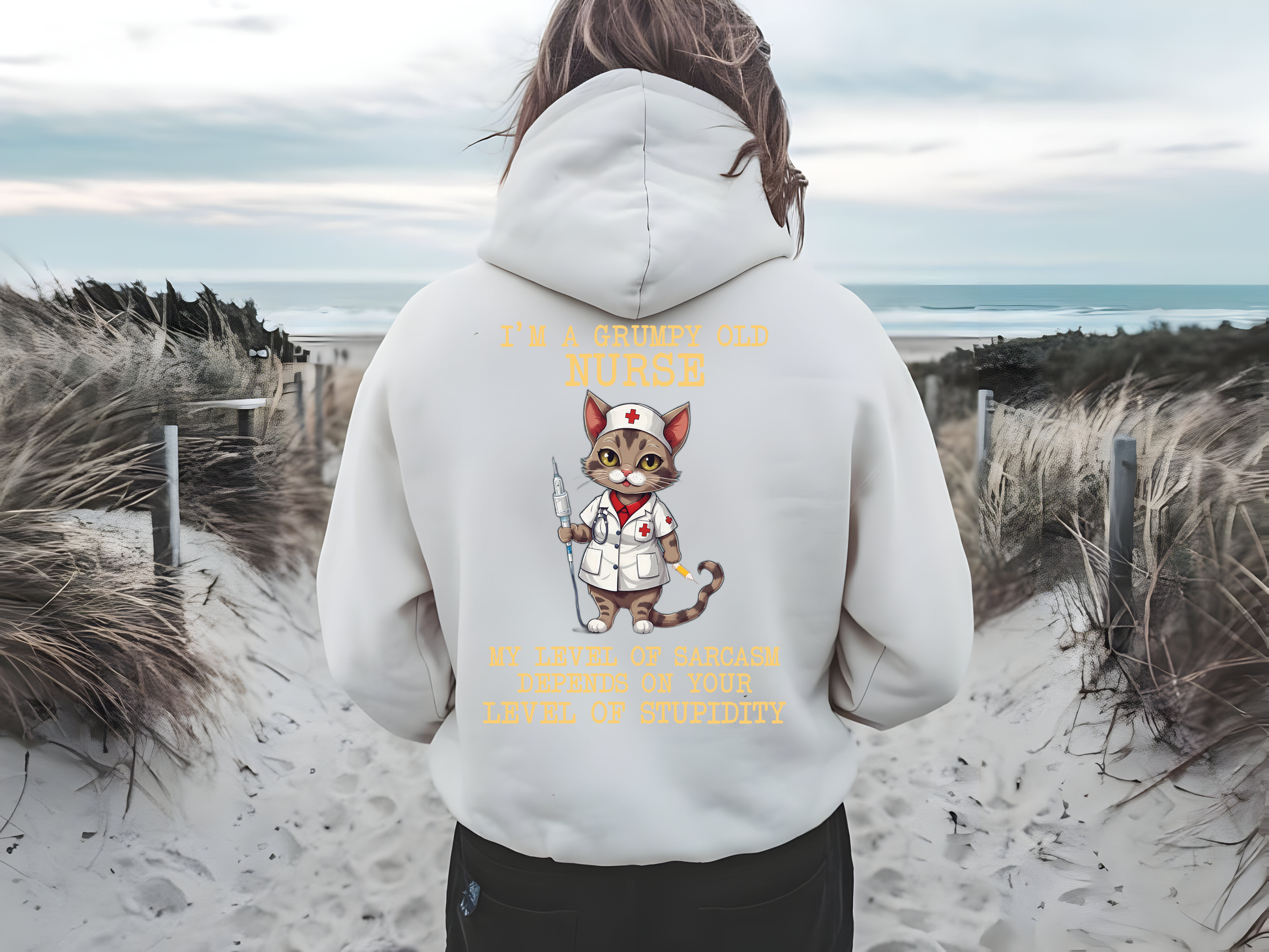 A woman wearing a cozy white hoodie with a humorous design featuring a cartoon cat dressed as a nurse. The back of the hoodie reads "I'M A GRUMPY OLD NURSE" with a playful graphic of the cat holding a syringe, while the quote "MY LEVEL OF SARCASM DEPENDS ON YOUR LEVEL OF STUPIDITY" is displayed prominently. Set against a scenic beach backdrop, this funny hoodie is perfect for healthcare workers and makes a great gift for nurses who appreciate lighthearted and comfortable apparel.