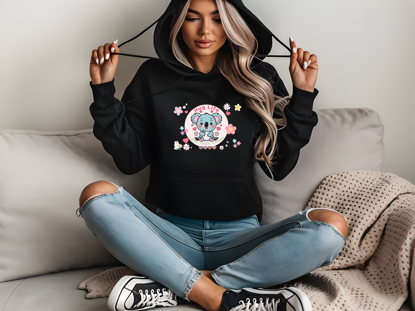 Stylish young woman sitting casually on a couch wearing a black hoodie with a cute koala design featuring the text 'Hug Life,' surrounded by flowers, hearts, and stars, perfect for cozy and trendy vibes.