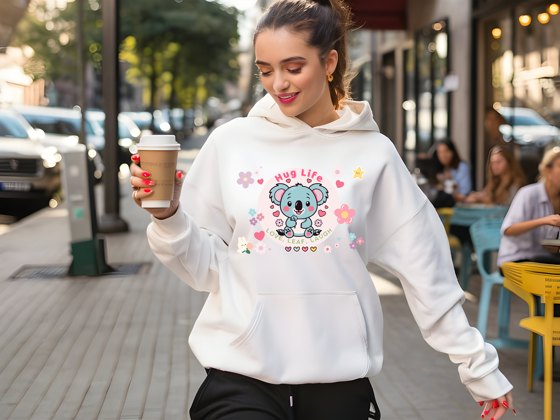 Chic woman walking outdoors wearing a white hoodie with a playful 'Hug Life' koala design, holding a coffee cup, and exuding casual, cheerful streetwear vibes.